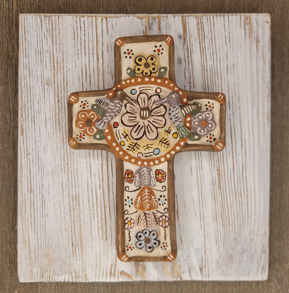 A hand-painted wooden cross plaque with intricate folk art patterns, displayed on a distressed white wooden board over a rustic brown background.