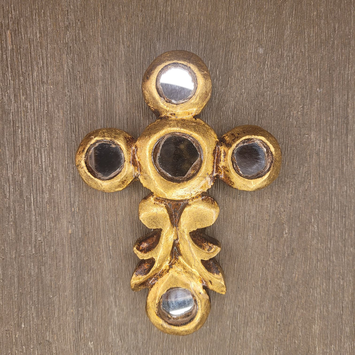 A gold-toned ornate cross-shaped wall hook or decorative hardware piece with circular recessed details, shown against a textured brown wood background.