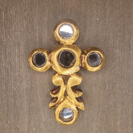 A gold-toned ornate cross-shaped wall hook or decorative hardware piece with circular recessed details, shown against a textured brown wood background.